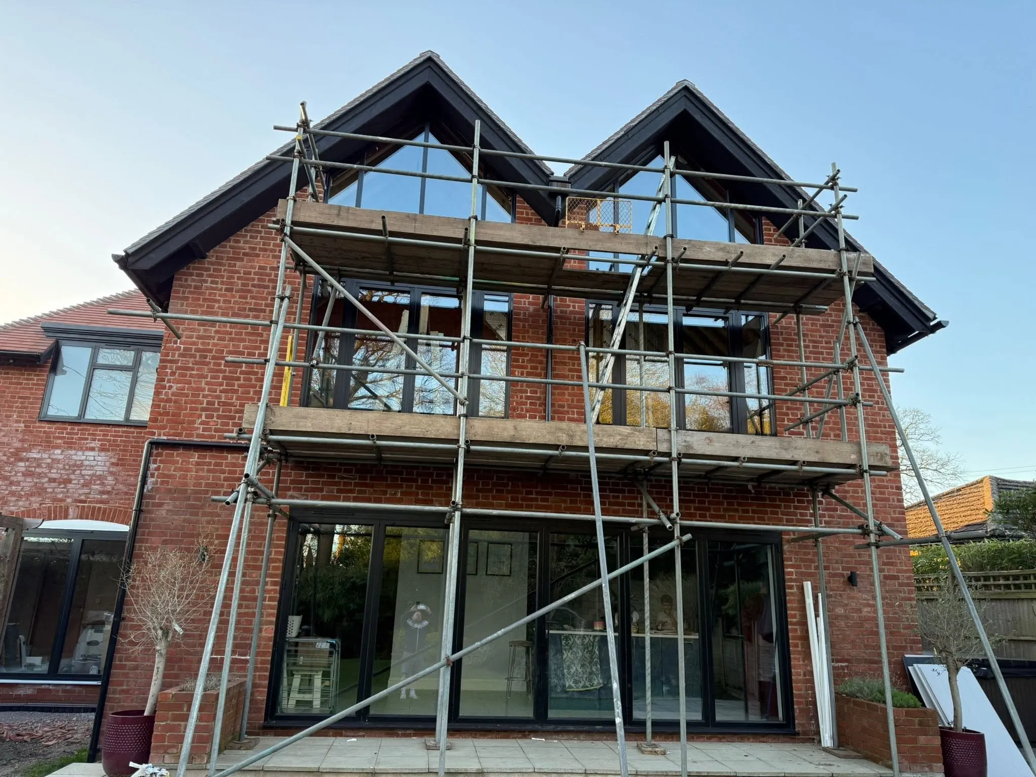 Rear of a brick house with new black aluminium gable windows, first-floor doors and wide glazed doors below, with scaffolding still in place