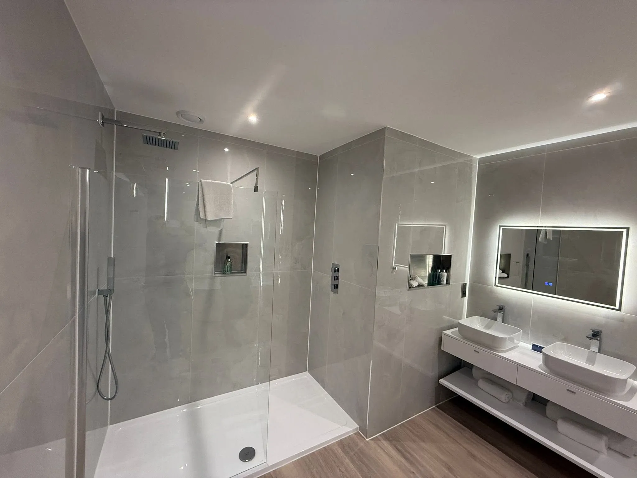 Bathroom with a walk-in shower, twin basins on a floating shelf and a backlit mirror