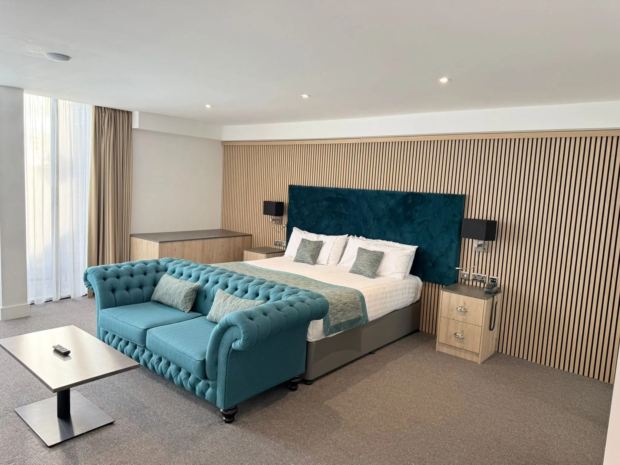 Hotel suite with a teal buttoned headboard and sofa, a slatted timber feature wall and full-height windows